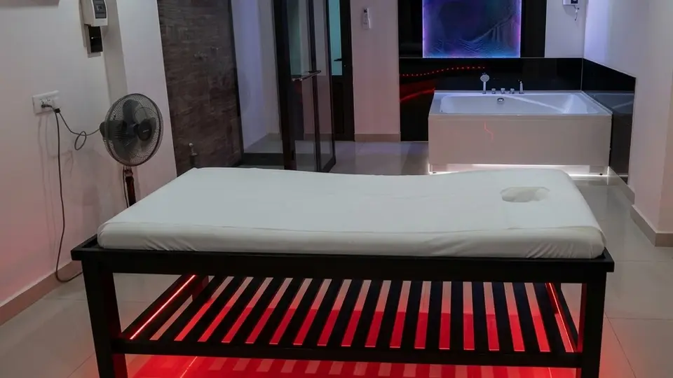 Interior of a room featuring a massage bed with red LED under-lighting and a large jacuzzi bathtub in the background